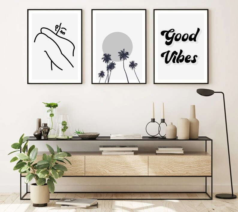 May include: Three framed art prints. The left print features a minimalist line drawing of a person. The center print shows a gray sun and palm trees. The right print has the words "Good Vibes" in a black, retro font.