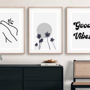 May include: Three framed art prints with minimalist designs. One features a line drawing of a person, another a sun and palm trees, and the third says "Good Vibes" in black script. The frames are light brown.