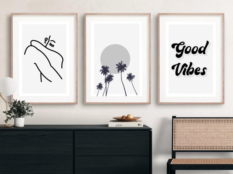 May include: Three framed art prints with minimalist designs. One features a line drawing of a person, another a sun and palm trees, and the third says "Good Vibes" in black script. The frames are light brown.