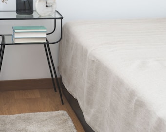 Könnte beinhalten: Ein weißes Bett mit einem beigefarbenen Leinen-Bettbezug und einem schwarzen Metall-Beistelltisch mit einer weißen Uhr und Büchern. Ein hellbrauner Teppich liegt auf dem Boden.