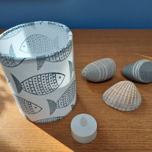 May include: A fabric lampshade with a white background and a pattern of grey fish. Beside it are three grey stones, a seashell, and a small white candle. The lampshade is cylindrical.