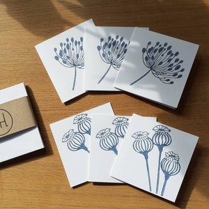 May include: A set of white square cards featuring blue floral designs, including seed heads and delicate blooms. A white box with a brown band displaying the initials "AH" is also present, along with a pen. The cards are arranged on a wooden surface.
