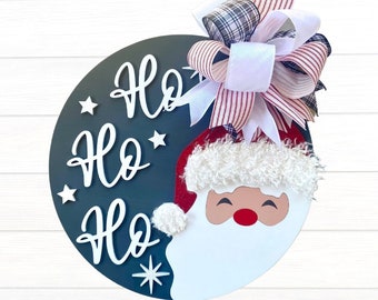 May include: A black and white wooden round sign with the words "Ho Ho Ho" in white lettering. A Santa Claus face with a white beard and red hat is on the right side of the sign. A red and white plaid bow with a white ribbon is on top of the sign.