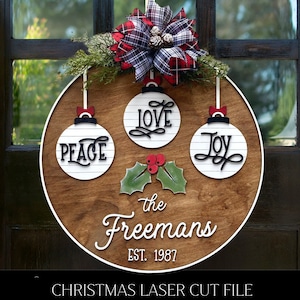 May include: A wooden Christmas ornament with a white background and black lettering. The ornament features the words "Peace", "Love", and "Joy" on three separate ornaments. The ornament also includes the text "The Freemans Est. 1987".
