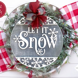Może przedstawiać: Okrągły, szary drewniany znak z napisem "Let It Snow" białymi literami. Znak jest ozdobiony białymi płatkami śniegu i otoczony zielonym wieńcem z czerwonymi jagodami. Czerwono-biała szachownica znajduje się po lewej stronie obrazu.