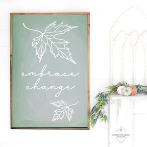 May include: A green wooden sign with a white outline of a maple leaf and the text "embrace change". The sign is hanging on a white wall with a garland of greenery and white pumpkins.