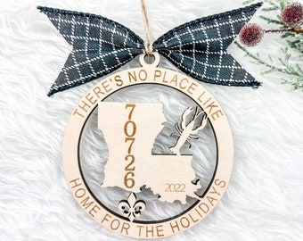 May include: A wooden Christmas ornament with a laser-cut design of the state of Louisiana, the text "There's no place like home for the holidays", and the area code "70726".