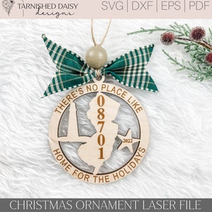 May include: A wooden Christmas ornament shaped like the state of New Jersey with the text "There's no place like home for the holidays" and the numbers "08701" inside the state outline. The ornament has a green and white plaid bow attached to the top.
