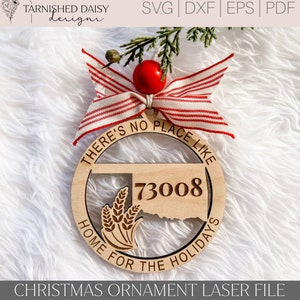 Könnte beinhalten: Ein Weihnachtsbaumschmuck aus Holz mit einem rot-weiß gestreiften Band. Der Schmuck ist kreisförmig und trägt den Text "There's no place like home for the holidays" und die Nummer "73008" im Inneren. Der Schmuck zeigt auch eine Silhouette des Bundesstaates Oklahoma mit Weizenähren.