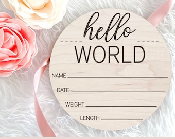 May include: A round wooden sign with the text "hello world" in a cursive font. The sign has blank lines for name, date, weight, and length. The sign is on a white fuzzy surface with pink flowers.