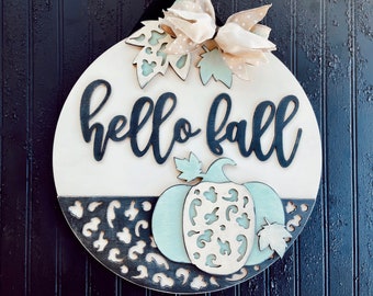 May include: A round wooden door hanger with a black and white leopard print design. The top half of the door hanger is white with the words "hello fall" in black lettering. A light blue pumpkin with a white cutout design is in the bottom half of the door hanger.