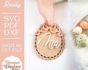May include: A wooden Easter egg ornament with a scalloped edge and a pink bow. The name "Mia" is written in white on the ornament. The ornament is on a white background with a pink ribbon and some speckled eggs.