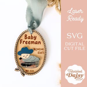 May include: Wooden oval ornament with a cowboy theme. The ornament features the text "Baby Freeman" and "Wrangling Hearts". A baby in a cowboy hat is in a bassinet. The text "May 2026" is also included.