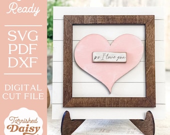 May include: A pink heart-shaped wooden cutout with the text "ps. I love you" on a small wooden sign inside a brown wooden frame. The frame is on a white wooden stand.