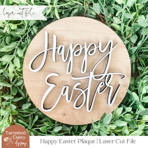 May include: A wooden plaque with the words "Happy Easter" in white script on a natural wood background. The plaque is surrounded by green foliage.
