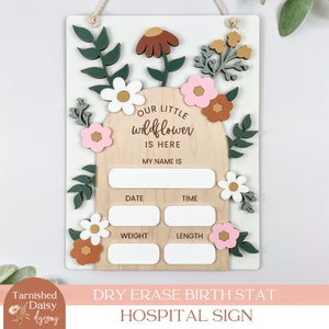 May include: A white wooden sign with a floral design and the text "Our Little Wildflower is Here" and spaces to write in the baby's name, date, time, weight, and length. The sign is a dry erase birth stat hospital sign.