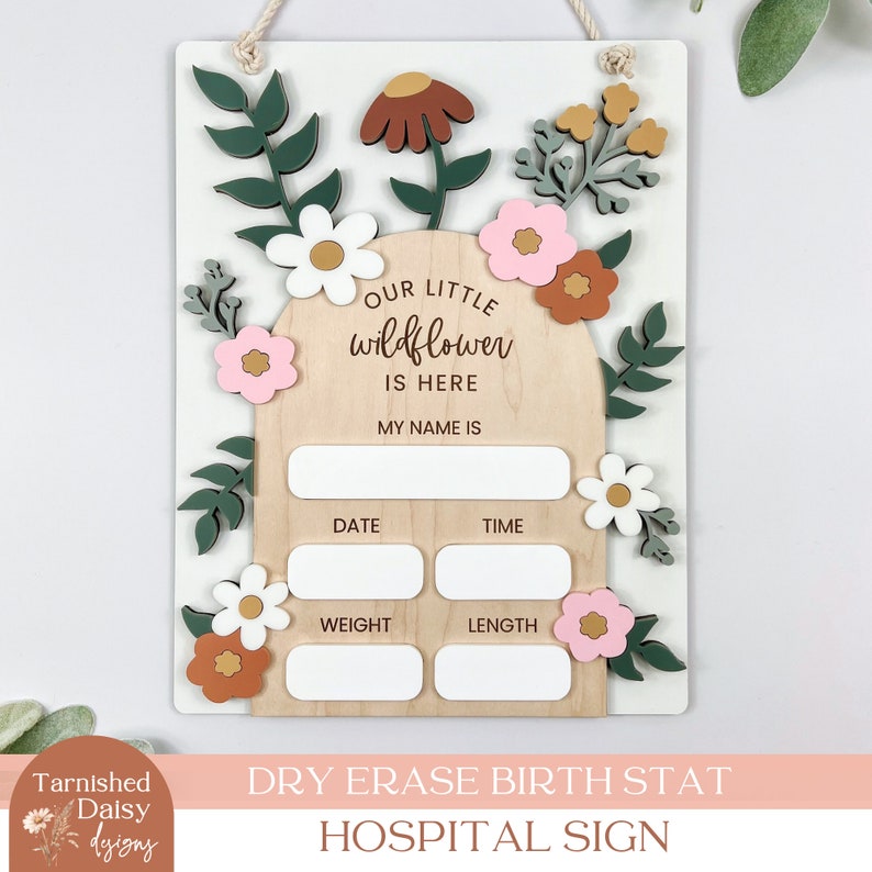 May include: A white wooden sign with a floral design and the text "Our Little Wildflower is Here" and spaces to write in the baby's name, date, time, weight, and length. The sign is a dry erase birth stat hospital sign.
