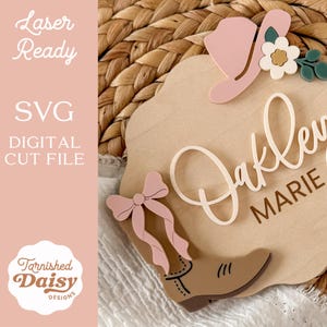 K&ouml;nnte beinhalten: Ein Holzschild mit einem Cowboy-Stiefel, Hut und floralen Akzenten. Der Name "Oakley Marie" ist in einer Schreibschrift dargestellt. Das Schild enth&auml;lt auch den Text "Laser Ready", "SVG DIGITAL CUT FILE" und das Logo "Tarnished Daisy Designs".