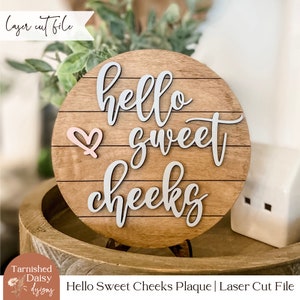 Puede incluir: Una placa de madera con el texto "hello sweet cheeks" en una fuente blanca de escritura. Un corazón rosa está en la esquina superior izquierda de la placa. La placa está en un soporte de madera.