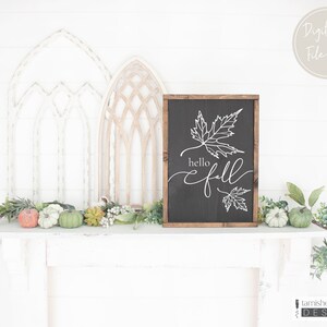 May include: A black chalkboard sign with a wooden frame, featuring a white outline of two maple leaves and the text "hello fall". The sign is sitting on a white mantel with a garland of greenery and small pumpkins.