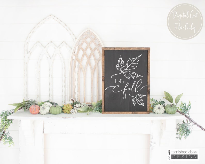 May include: A black chalkboard sign with a wooden frame, featuring a white outline of two maple leaves and the text "hello fall". The sign is sitting on a white mantel with a garland of greenery and small pumpkins.