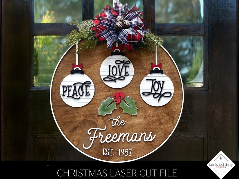 May include: A wooden Christmas ornament with a white background and black lettering. The ornament features the words "Peace", "Love", and "Joy" on three separate ornaments. The ornament also includes the text "The Freemans Est. 1987".