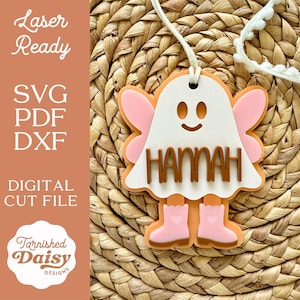 May include: A layered, laser-cut ghost-shaped pendant with the name "HANNAH" in brown letters. The ghost has pink wings and boots, with a white body and orange outline. The pendant hangs from a white cord. Text on the left side reads "Laser Ready" and "Digital Cut File."