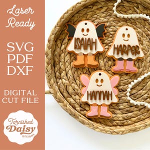 Puede incluir: Tres adornos de madera con forma de fantasma con nombres: Isaiah, Harper y Hannah. Cada fantasma tiene una cara sonriente, alas y botas. Los adornos están sobre una cesta tejida. El texto "Laser Ready", "SVG PDF DXF" y "Digital Cut File" es visible.