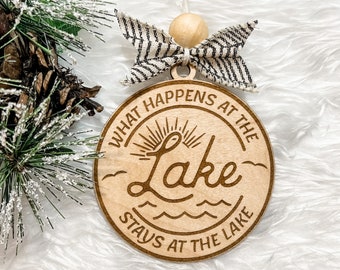 May include: A wooden ornament with the text "What happens at the lake stays at the lake" and a sun with a lake wave design. The ornament is decorated with a black and white striped bow.