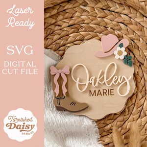 K&ouml;nnte beinhalten: Namensschild aus Holz mit Cowboy-Motiv. Verf&uuml;gt &uuml;ber eine hellbraune Holzbasis mit dem Namen "Oakley Marie" in kursiver Schrift. Verziert mit einem rosa Cowboyhut, einer Schleife, einem Cowboy-Stiefel und einer Blume. Das Schild enth&auml;lt auch den Text "Laser Ready" und "SVG Digital Cut File".