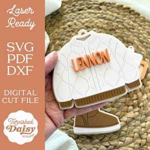 May include: A white sweater-shaped ornament with the name "Lennon" in orange letters. The ornament has brown trim and is paired with a brown boot-shaped ornament. The image also includes text that says "Laser Ready", "SVG PDF DXF", "Digital Cut File", and the logo for Tarnished Daisy Designs.