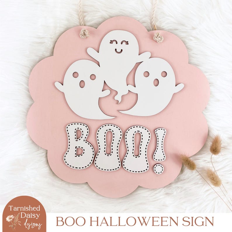 Może przedstawiać: R&oacute;żowe drewniane halloweenowe szyld z napisem "BOO!" białymi literami z obszytym konturem. Trzy białe wycięcia duch&oacute;w są rozmieszczone wok&oacute;ł napisu "BOO!"