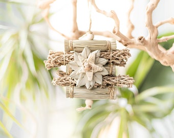 Può includere: Un ornamento a forma di stella intrecciato in modo naturale appeso a un ramo. L'ornamento è fatto di paglia e ha un aspetto rustico e terroso.