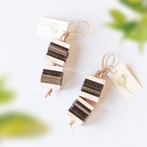 May include: Two bird toys made of wood and corrugated cardboard. Each toy has a series of stacked cardboard pieces held together by wooden dowels. A small tag with a bird illustration is attached to each toy with twine. The toys are designed for birds.
