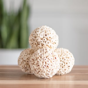 Puede incluir: Cuatro bolas decorativas blancas hechas de pequeñas piezas redondas blancas. Las bolas están apiladas en forma de pirámide sobre una superficie de madera.