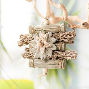 May include: A natural woven star-shaped ornament hangs from a branch. The ornament is made of straw and has a beige color. It is a decorative piece for a home or a bird's cage.