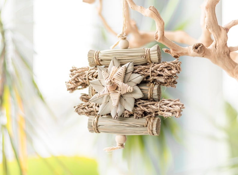 May include: A natural woven star-shaped ornament hangs from a branch. The ornament is made of straw and has a beige color. It is a decorative piece for a home or a bird's cage.