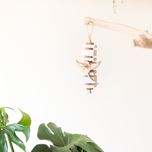 May include: A hanging wooden bird mobile. The mobile features a stack of white and natural wood blocks with a laser-cut bird in the center. It is suspended from a natural wood branch with a twine loop.