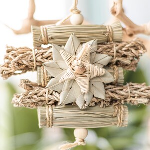 May include: A natural woven star ornament with a wooden bead hanging from the bottom. The star is surrounded by bundles of natural fibers, creating a rustic and organic look.