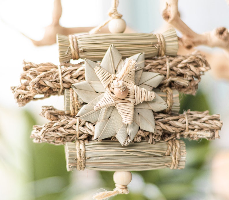 May include: A natural woven star ornament with a wooden bead hanging from the bottom. The star is surrounded by bundles of natural fibers, creating a rustic and organic look.
