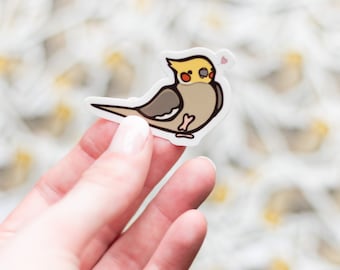 Puede incluir: Una pegatina de un cacatúa crestado amarillo con un corazón rosa sobre su cabeza. El cacatúa está mirando hacia la derecha y tiene un cuerpo marrón claro con un pecho blanco y una cresta amarilla. La pegatina está en una mano.