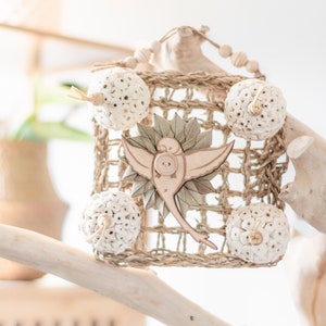May include: A decorative birdcage-shaped toy for birds, crafted from natural materials. The cage features a woven design, adorned with wooden beads, decorative spheres, and a wooden bird cutout with a button detail. The color palette is neutral, with beige and light brown tones.