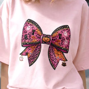 Pode incluir: Uma t-shirt de manga curta rosa claro com um grande gráfico central de um laço. O laço tem um padrão rosa, laranja e preto com manchas de leopardo, riscas de zebra, fantasmas, gatos pretos, abóboras e caveiras. Pequenos berloques pendem do laço.