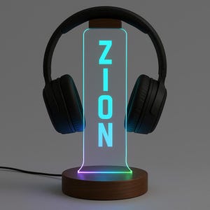 Può includere: Un supporto per cuffie nero con il nome "ZION" illuminato in blu. Il supporto ha una base in legno e un pannello in acrilico trasparente. Le cuffie sono nere e poggiano sulla parte superiore del supporto.