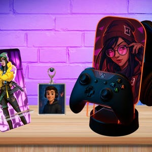Pode incluir: Uma coleção de acessórios de jogos com arte de personagens. Inclui um suporte para telefone, um chaveiro e um suporte para controlador com fones de ouvido. Os itens são exibidos em uma superfície de madeira contra uma parede de tijolos roxos.