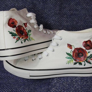 Op de afbeelding: Een paar witte high-top canvas schoenen met rode klaproosbloemontwerpen die erop zijn geschilderd.