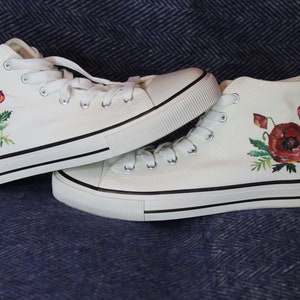 Op de afbeelding: Een paar witte hoge canvas sneakers met rode klaproos-borduurwerk.