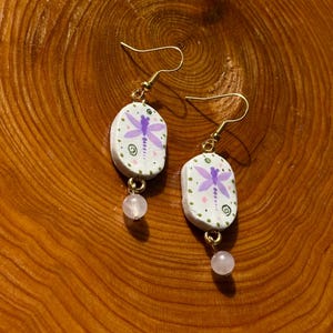 May include: Hand-painted earrings with a dragonfly design. The earrings feature a white base with a purple dragonfly, green dots, and a pink bead. The gold-colored hooks and the pink beads add a touch of elegance to these unique earrings.