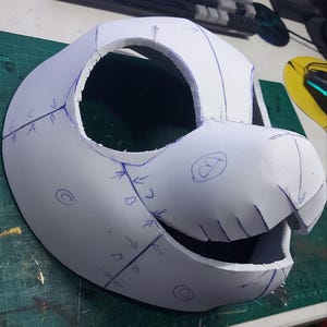 Op de afbeelding: Een witte schuimrubberen maskerbasis met een grote ronde oogopening en een uitstekende snuit. Het oppervlak is gemarkeerd met blauwe inktlijnen, pijlen en letters zoals A, B en C, die de montagepunten voor de constructie aangeven.