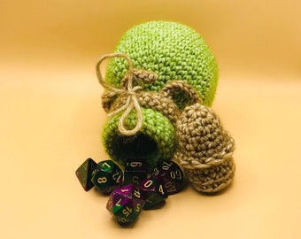 May include: A green crocheted dice bag with a drawstring closure, a small brown crocheted dice bag, and several multi-sided dice in shades of green, purple, and black. The dice are scattered around the bags.